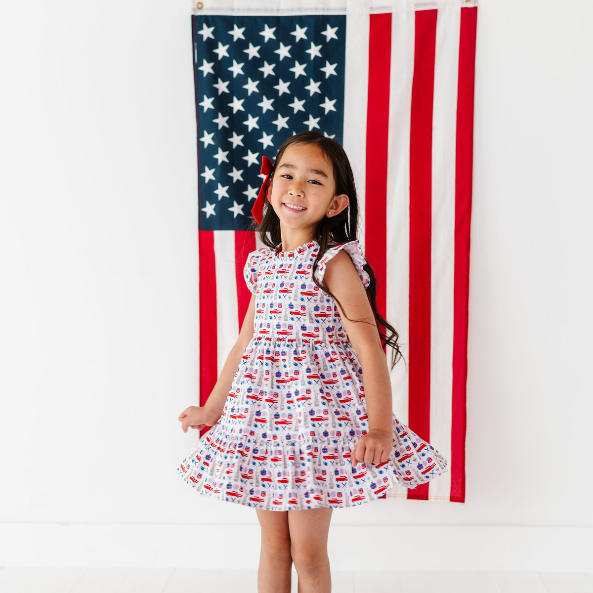 Young girl in a red, white, and blue Americana print dress standing in front of an American flag.