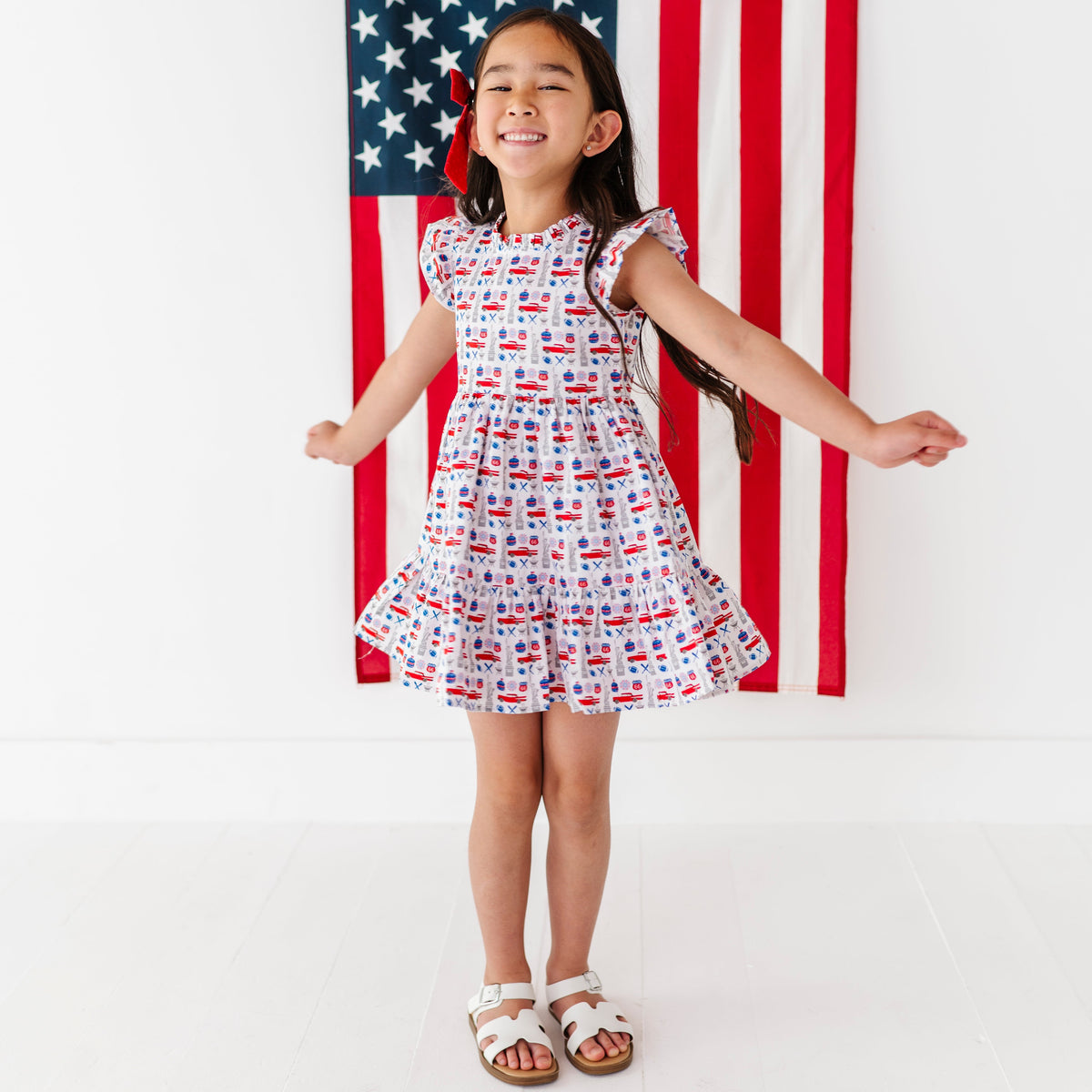 Young girl in a red, white, and blue Americana print dress standing in front of an American flag.