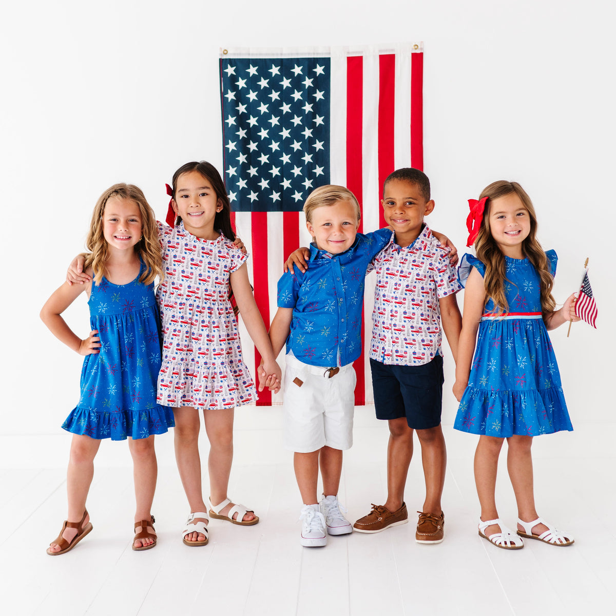 Five children in patriotic outfits standing in front of an American flag on a white background