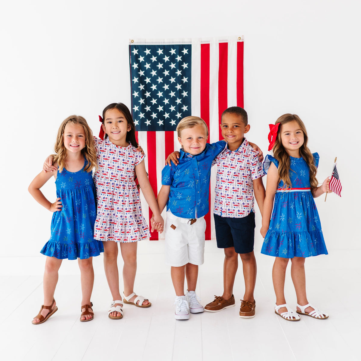 Five children in patriotic outfits standing in front of an American flag on a white background