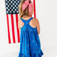 Young girl in a blue dress with ajuly 4th firework print standing in front of an American flag.
