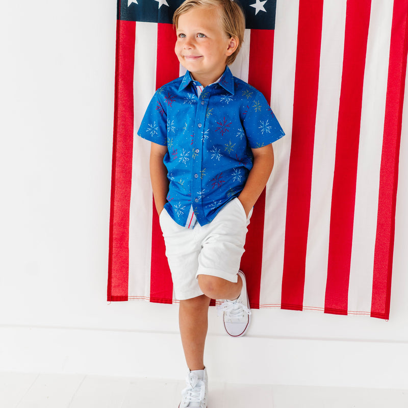 Child wearing a firework print shirt and white shorts standing in front of an American flag.