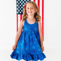 Young girl in a blue dress with ajuly 4th firework print standing in front of an American flag.