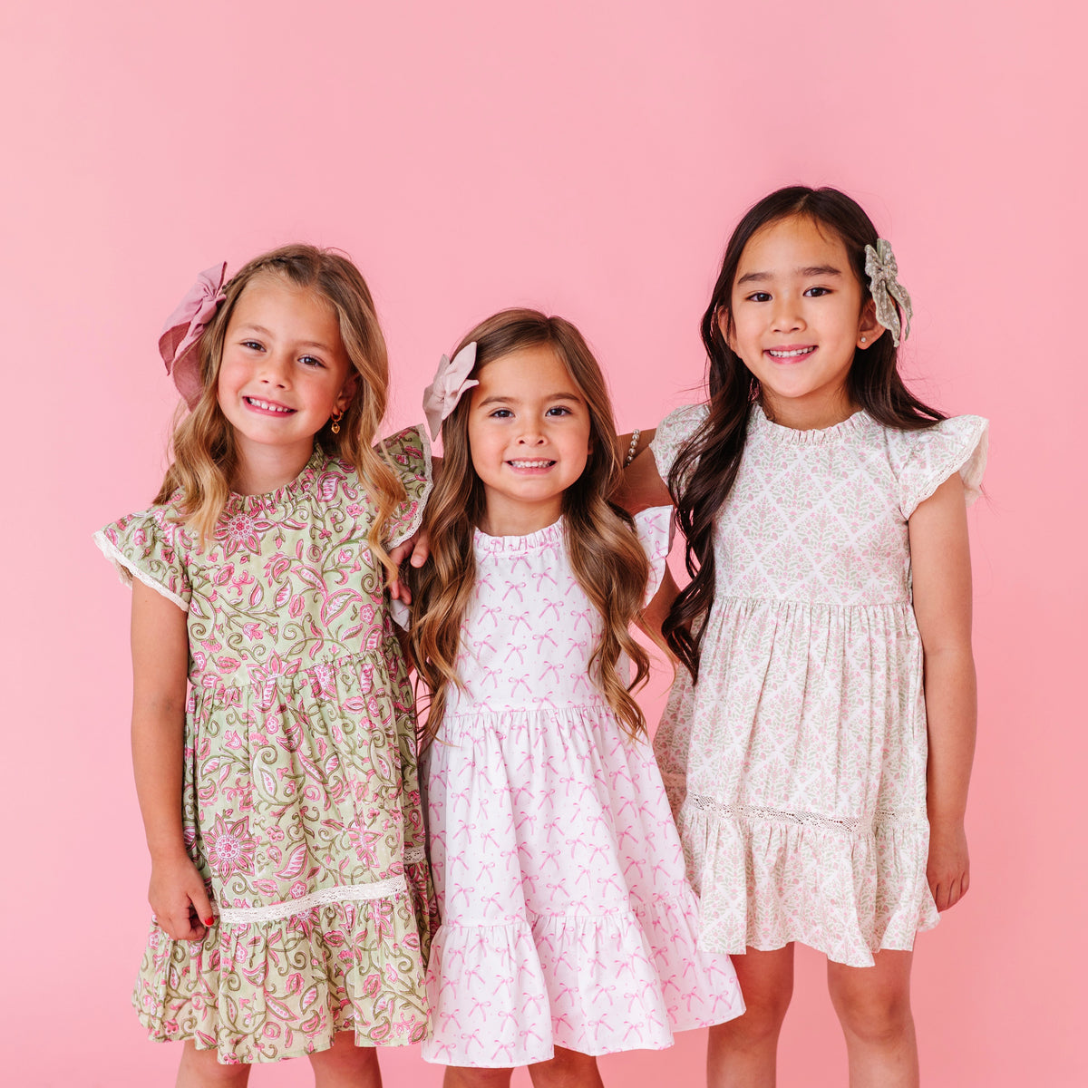 Three young girls wearing floral dresses against a pink background